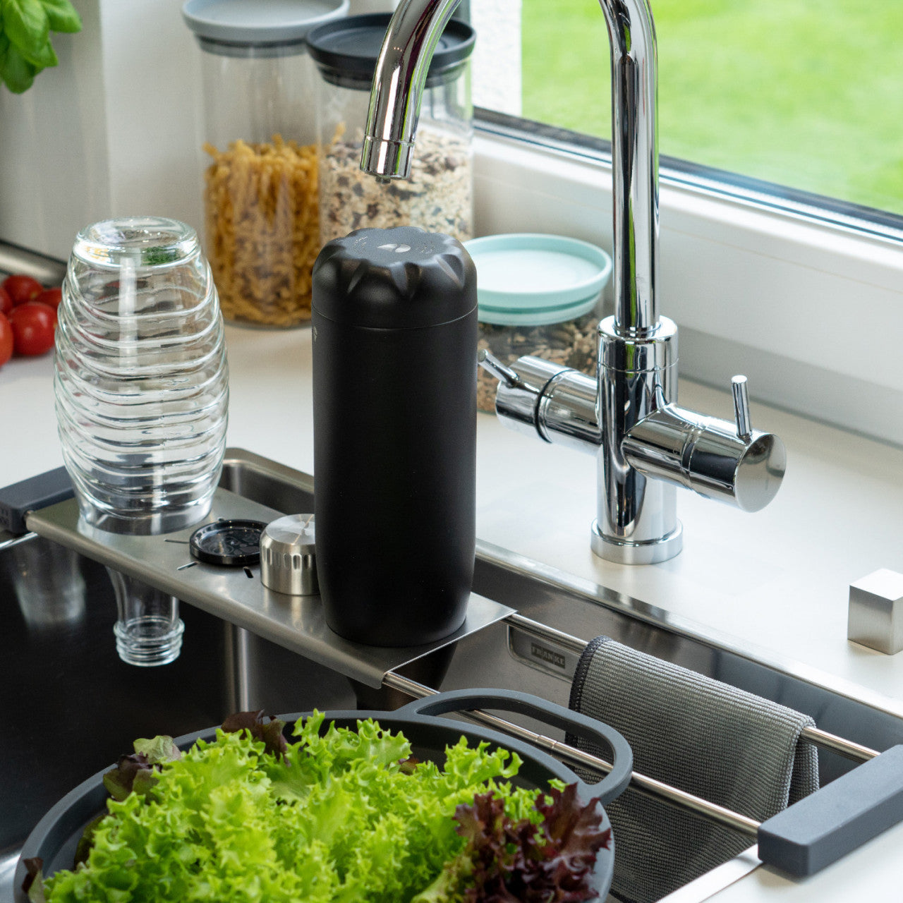 Eine Abtropfhilfe Drip.it steht neben einer Küchenspüle, im Hintergrund sind Salat, Glaswaren und Einmachgläser zu sehen.