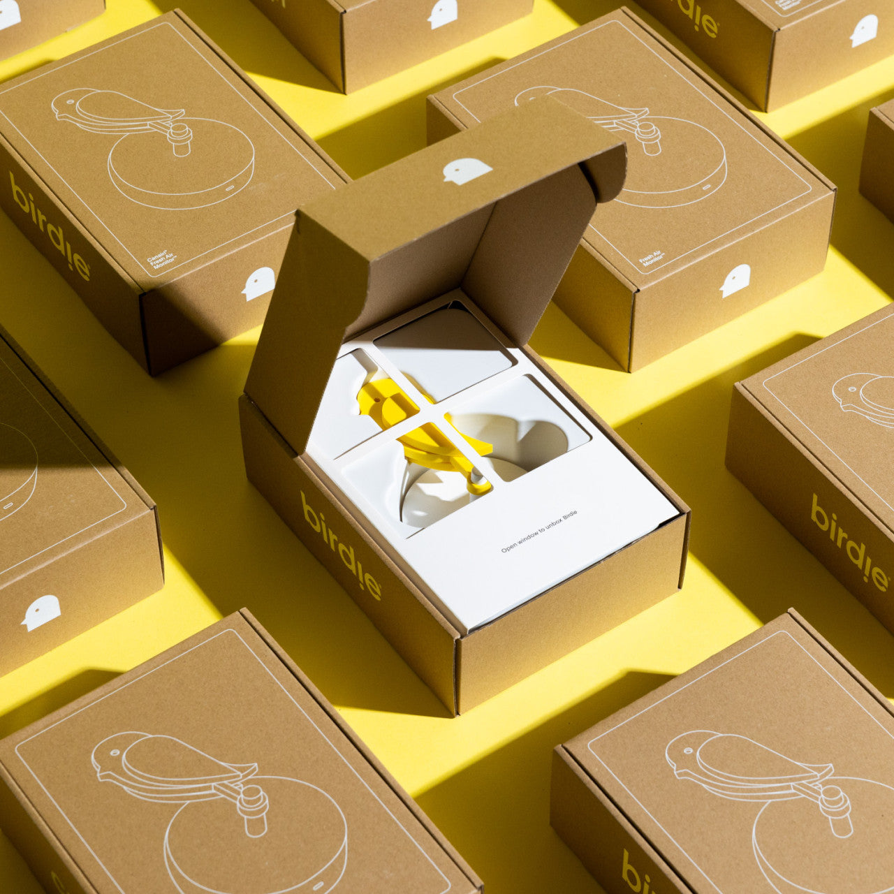In einer offenen Box befindet sich ein gelber Frischluftmonitor Birdie 2.0 mit CO2-Sensor, umgeben von gleichartigen geschlossenen Boxen auf gelbem Untergrund.