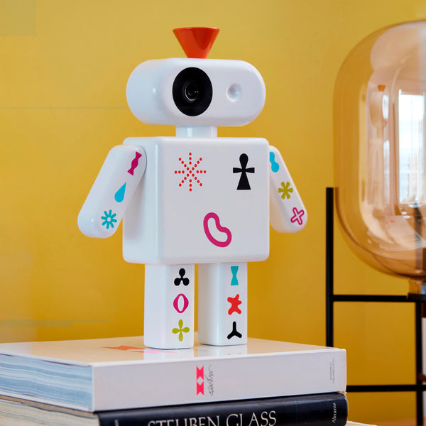 Ein weißer Spielzeugroboter mit bunten Symbolen steht auf gestapelten Büchern vor einer gelben Wand und einer Lampe.