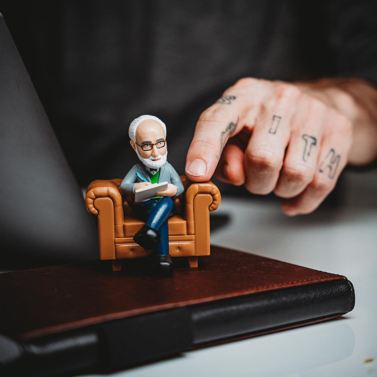 Eine Hand zeigt auf den Schreibtisch-Psychologen, eine kleine Figur eines Mannes mit Brille, der auf einem Stuhl sitzt und auf einem Notizbuch steht.