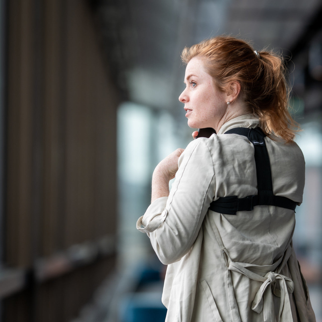 Eine Frau mit roten Haaren trägt einen beigen Mantel und den Haltungskorrektor Haltungstrainer schwarz M-L und schaut aus einem Fenster.