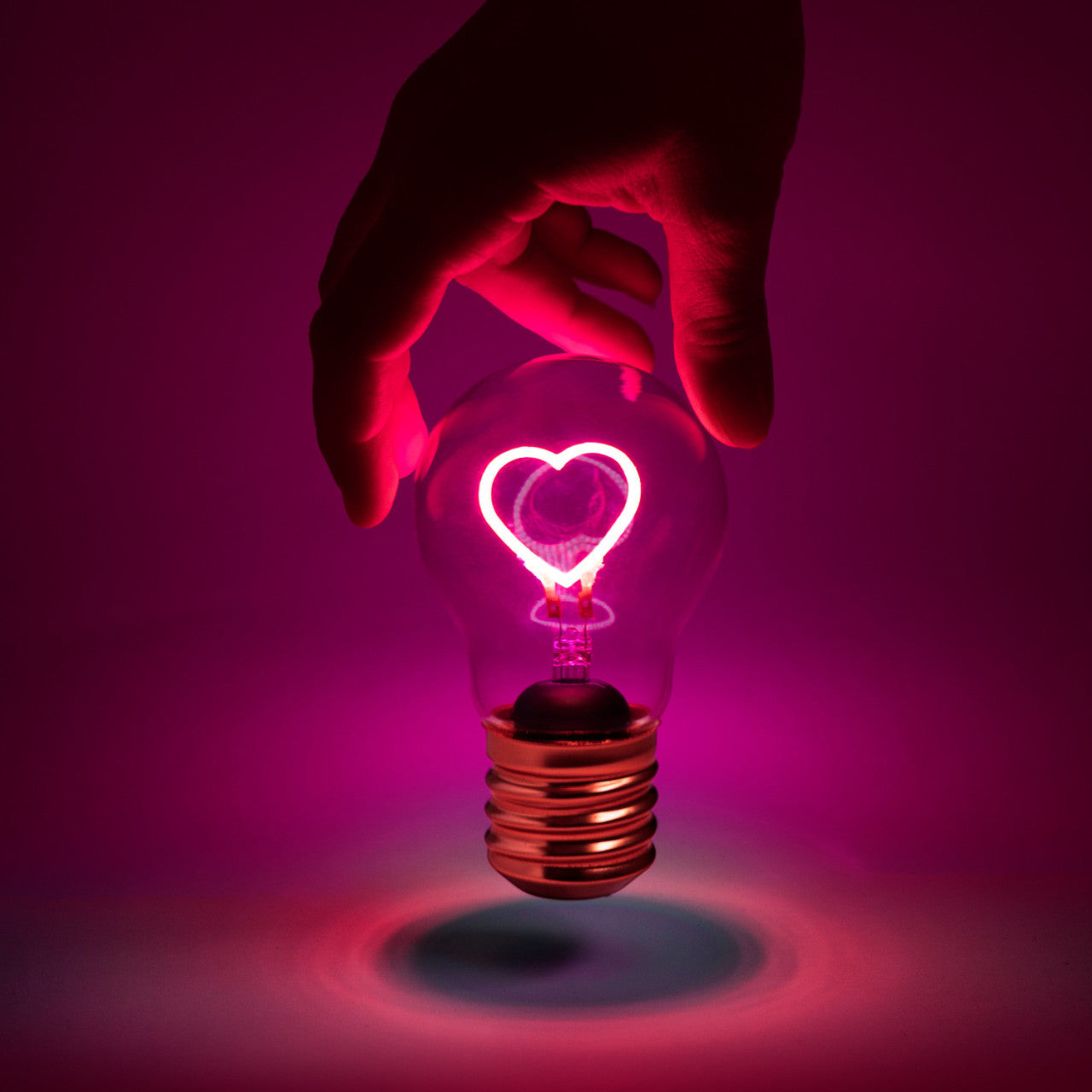 Eine Hand hält die Kabellose LED-Glühbirne - Herz mit einem leuchtend rosa herzförmigen Glühfaden vor einem violetten Hintergrund.