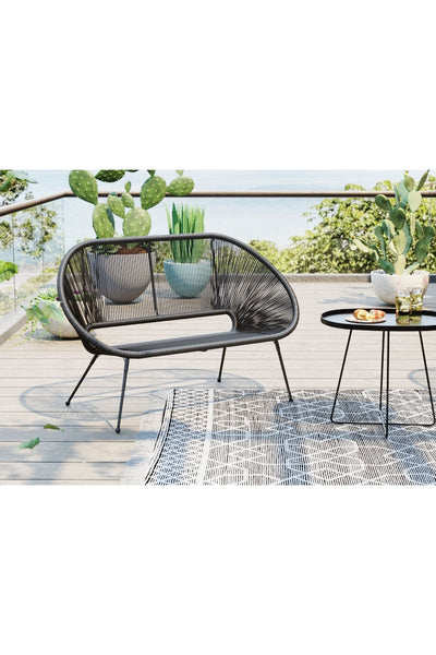 Ein schwarzer, moderner Outdoor-Liegesessel und ein runder Beistelltisch stehen auf einer Terrasse mit Teppich, umgeben von Grün und mit Blick auf das Wasser im Hintergrund.