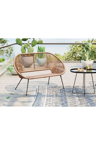 Ein brauner geflochtener Outdoor-Sofa und ein schwarzer runder Beistelltisch stehen auf einer Terrasse mit gemustertem Teppich, umgeben von Topfpflanzen und mit Blick auf Wasser und Grün.