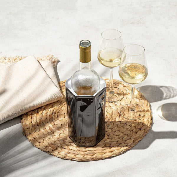 Eine Flasche Weißwein in einer Kühlbox mit zwei gefüllten Gläsern auf einer gewebten Matte neben einem gefalteten beigen Tuch.