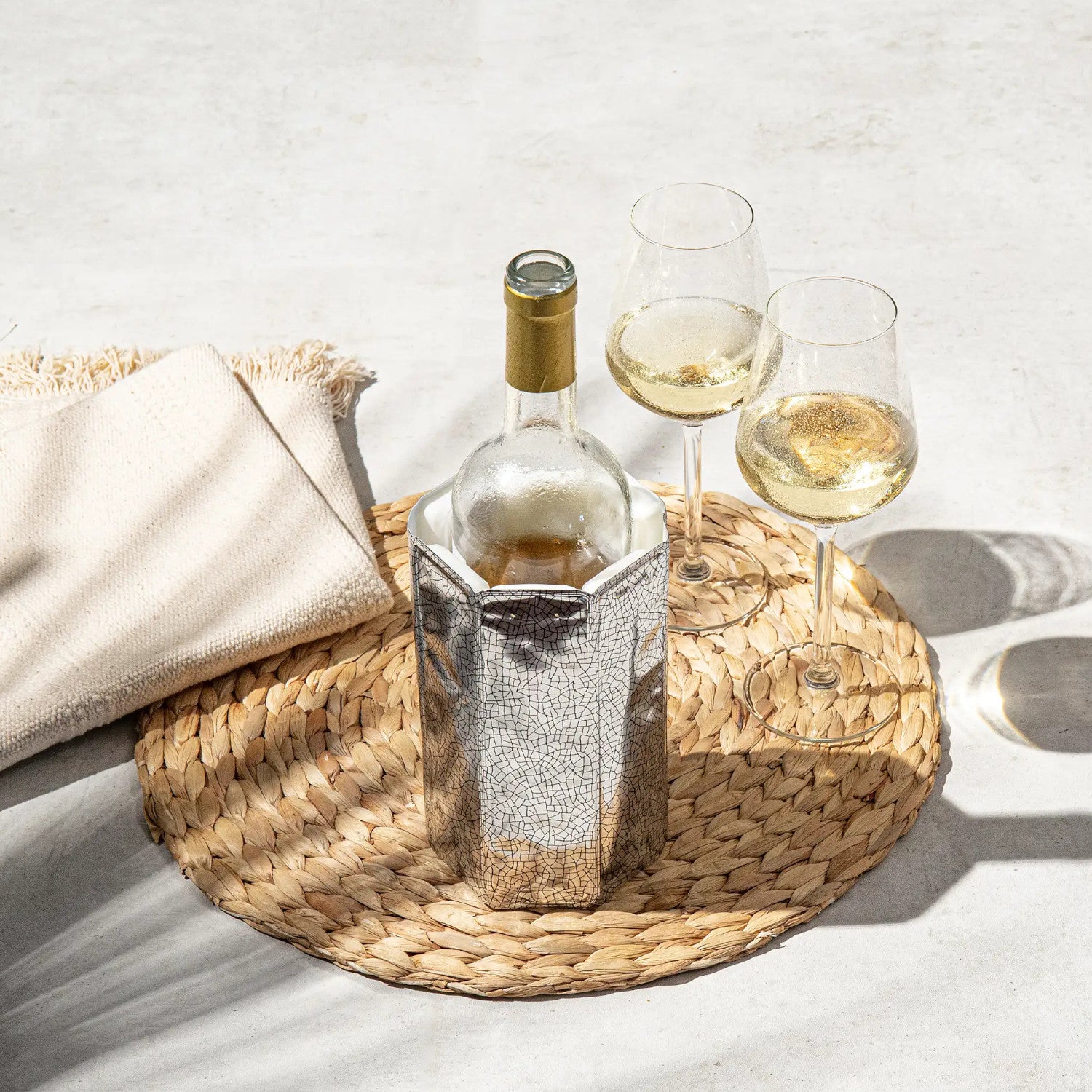 Gekühlte Weißweinflasche und zwei Gläser auf einer gewebten Matte mit einem beigen Tuch daneben.