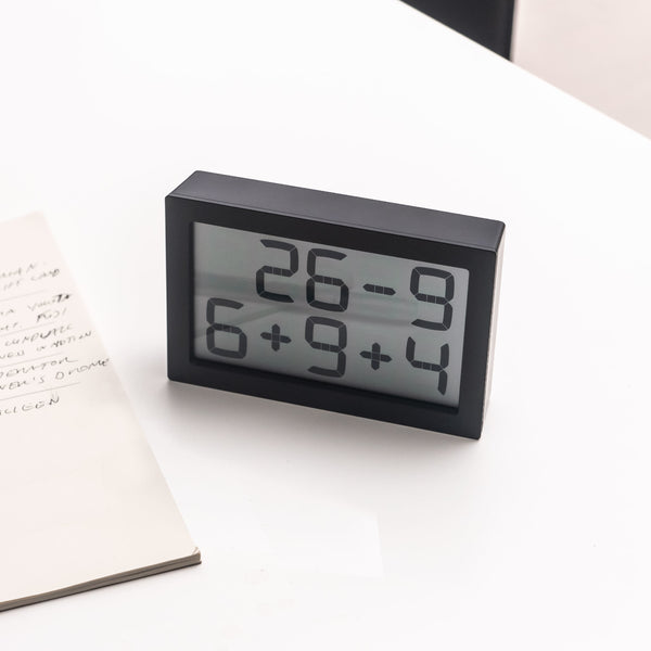 Eine Digitaluhr, die oben "26-9" und unten "6+9+4" anzeigt, auf einem weißen Schreibtisch neben einem Notizbuch.