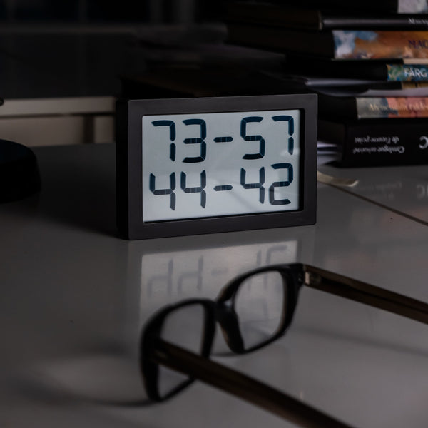 Digitaluhr mit Zahlenanzeige auf einem Schreibtisch, mit Brille und gestapelten Büchern im Hintergrund.