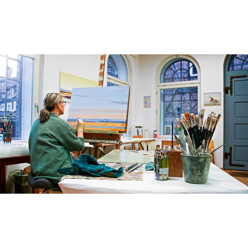 Eine Frau malt in einem hellen Atelier mit Pinseln auf dem Tisch ein Meeresbild, inspiriert von Anja Strucks Kunstwerk „Die Liebe zum Meer“ (2023).