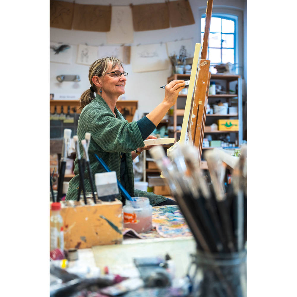 Eine Frau lächelt, während sie in einem hellen, mit Kunstwerken gefüllten Atelier an einer Staffelei malt. Im Vordergrund ist Anja Strucks „Die Liebe zum Meer“ (2023) zu sehen, mit Pinseln.