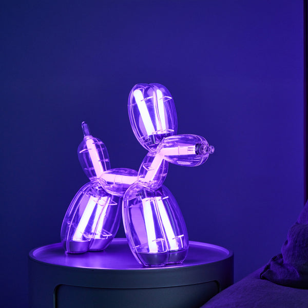 Eine Lampe in Form eines Ballonhundes leuchtet mit violettem Licht auf einem runden Tisch in einem schwach beleuchteten Raum.