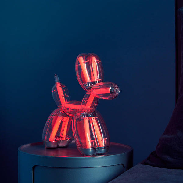 Eine durchsichtige Ballon-Hundelampe leuchtet rot auf einem runden Tisch vor einem dunkelblauen Hintergrund.