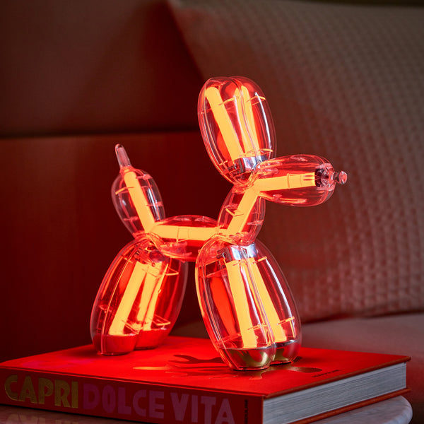 Eine rot leuchtende Ballonhund-Lampe sitzt auf einem "CAPRI DOLCE VITA"-Buch neben einem Bett mit einem strukturierten Kissen.