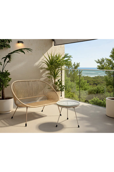 Eine moderne Terrasse mit einem Liegesessel aus Korbgeflecht, einem kleinen runden Tisch, Topfpflanzen und einem Glasgeländer mit Blick auf Bäume und das Meer im Hintergrund.