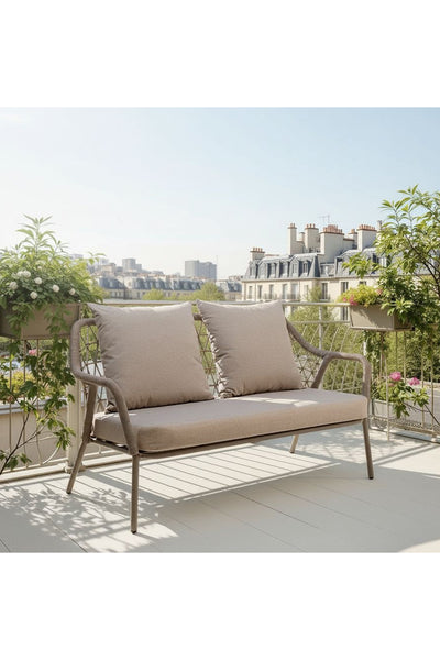 Ein beigefarbenes, gepolstertes Outdoor-Sofa steht auf einem Balkon mit Topfpflanzen und bietet einen Blick auf die Gebäude der Stadt bei klarem Himmel.