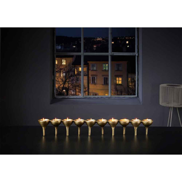 Acht brennende Kerzen in goldenen Haltern auf einem Tisch mit einer Stadtlandschaft, die durch ein Fenster bei Nacht zu sehen ist.