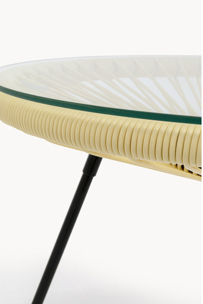 Nahaufnahme eines runden Tisches mit Glasplatte, beigefarbener geflochtener Rattankante und schwarzen Metallbeinen vor einem weißen Hintergrund.