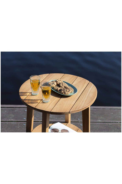 Ein runder Holztisch mit zwei Gläsern mit bernsteinfarbener Flüssigkeit und einem Teller mit Keksen steht im Freien in der Nähe von Wasser.