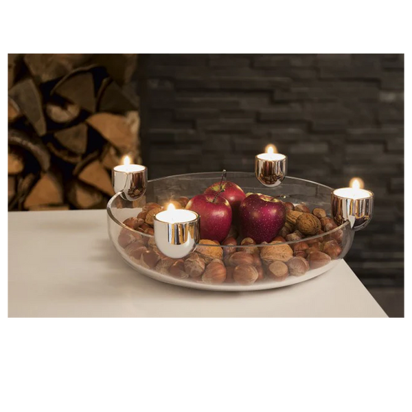 Glasschale mit Äpfeln und Nüssen, umgeben von fünf brennenden Kerzen in silbernen Haltern auf einer weißen Fläche.