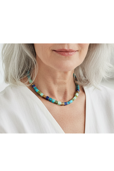 Petra Waszak: Collier „Bergsee“ besteht aus quadratischen blauen, grünen und goldenen Perlen mit einem goldenen Verschluss und einer verstellbaren Kette. Man sieht es hier getragen an dem Hals einer älteren Dame. 