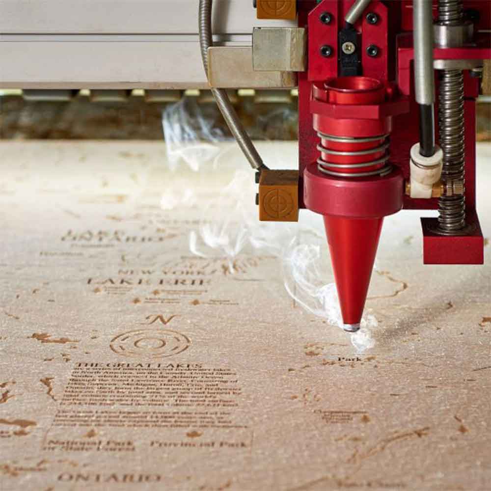 Eine Lasergravurmaschine, die detaillierten Kartentext und Grafiken auf eine flache Holzoberfläche ätzt, wobei kleine Rauchschwaden sichtbar sind.