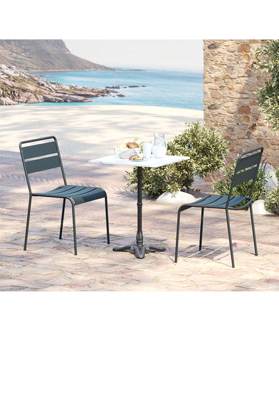 Zwei Metallstühle und ein runder Tisch auf einer Steinterrasse in Strandnähe, mit Speisen und Getränken auf dem Tisch und einer Küstenlandschaft im Hintergrund.