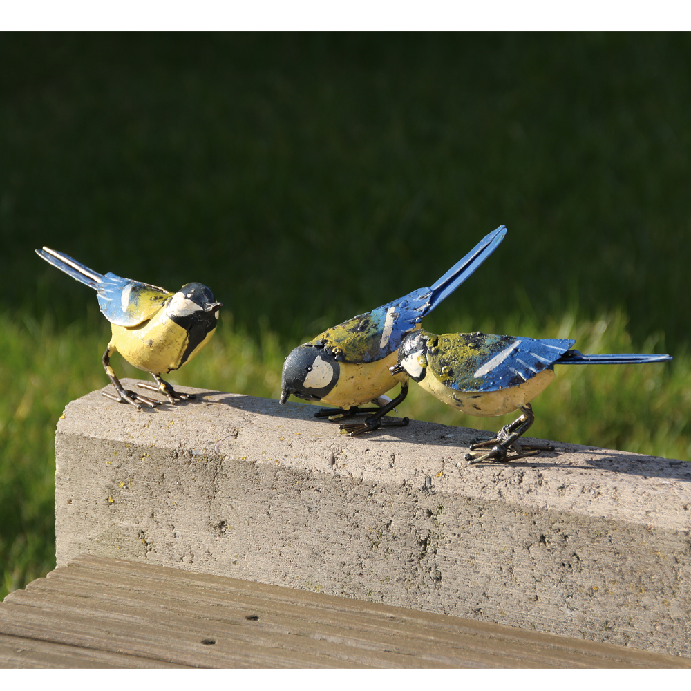 Drei blaue und gelbe Vogelskulpturen aus Metall sitzen im Sonnenlicht auf einem Zementvorsprung.