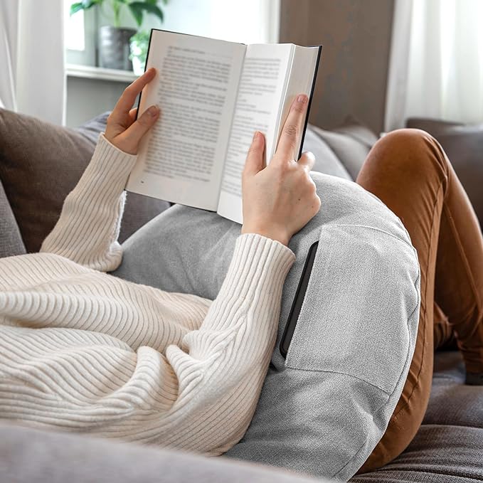 Eine Person in einem weißen Pullover liegt mit gekreuzten Beinen auf einem Sofa und liest bequem mit dem Lesekissen Classic mit Tasche – Grau, einem weichen Lesekissen aus deutscher Herstellung.