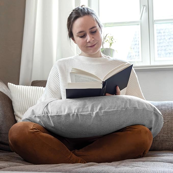 Eine Frau sitzt im Schneidersitz auf einem Sofa, liest mit Komfort und dem Lesekissen Classic mit Tasche - Grau auf ihrem Schoß und genießt einen ruhigen Moment am Fenster. Hergestellt in Deutschland.