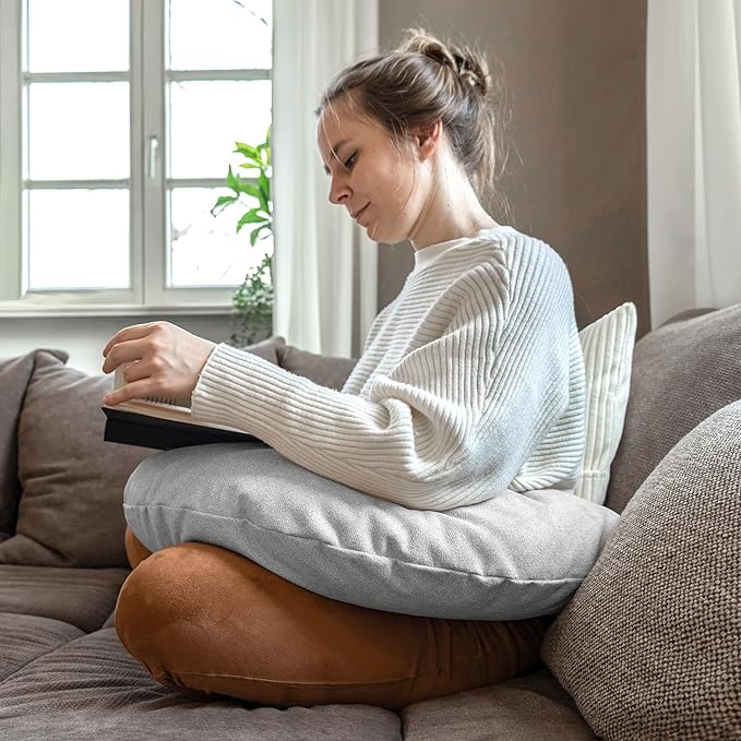 Eine Frau sitzt im Schneidersitz auf einem Sofa und liest. Das Lesekissen Classic mit Tasche – Grau liegt auf ihrem Schoß für optimalen Komfort. Hergestellt in Deutschland.