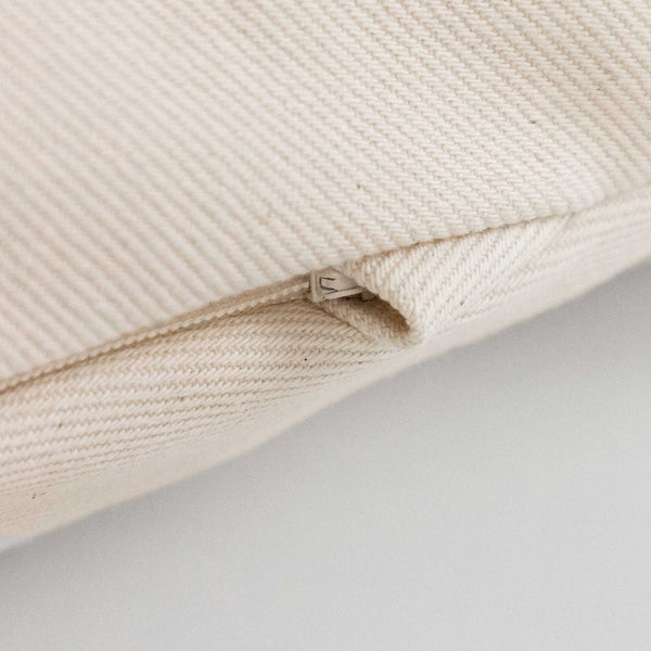 Nahaufnahme eines cremefarbenen Stoffkissens mit einem teilweise geöffneten Reißverschluss auf einer hellen Fläche.