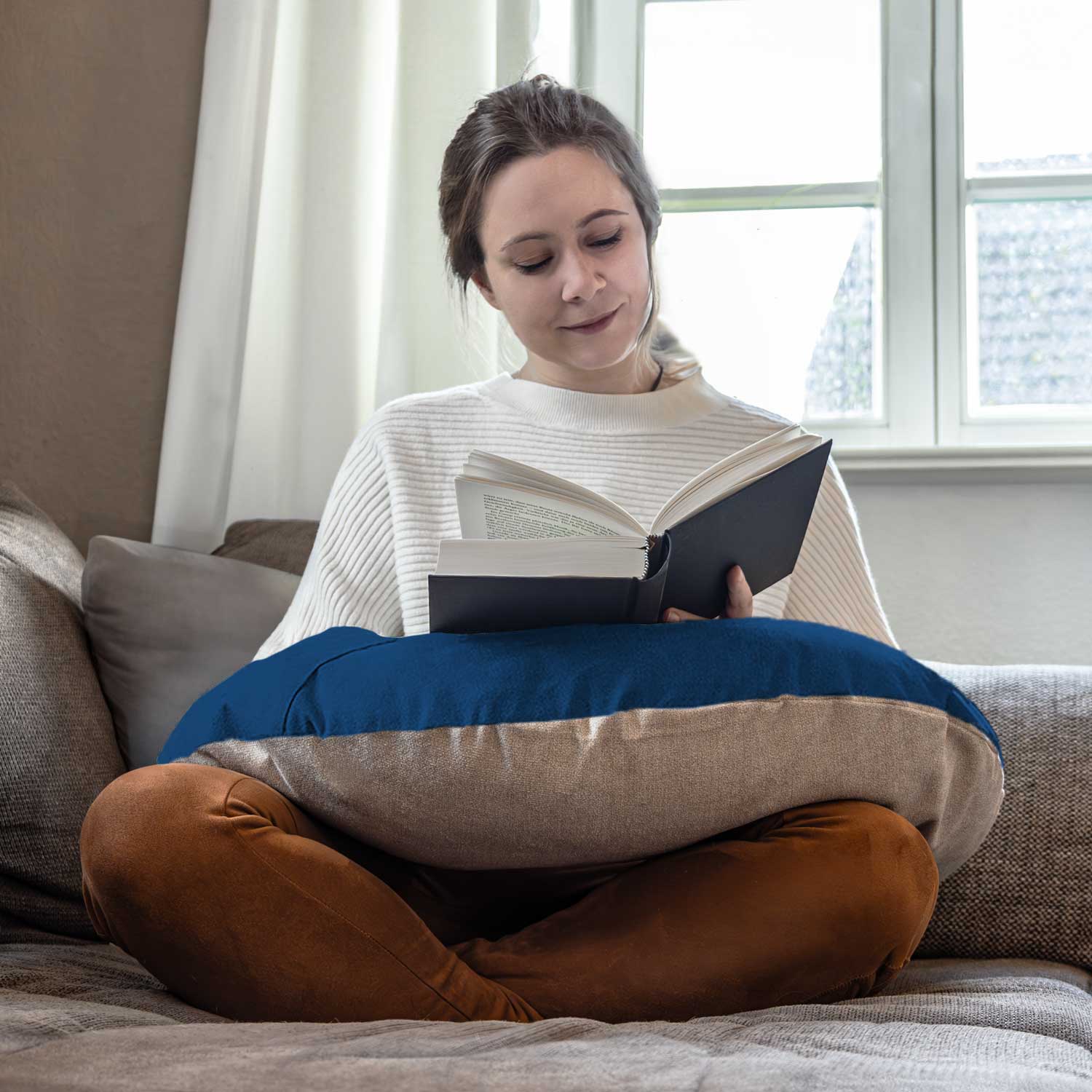 Frau sitzt im Schneidersitz auf einer Couch und liest ein Buch mit einem Kissen auf dem Schoß in der Nähe eines Fensters.