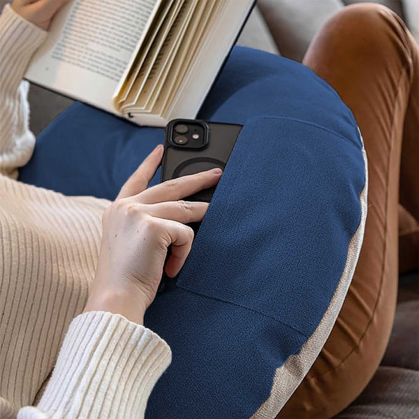 Eine Person sitzt mit einem blauen Kissen auf dem Schoß, hält ein Telefon in der Hand und liest ein Buch.