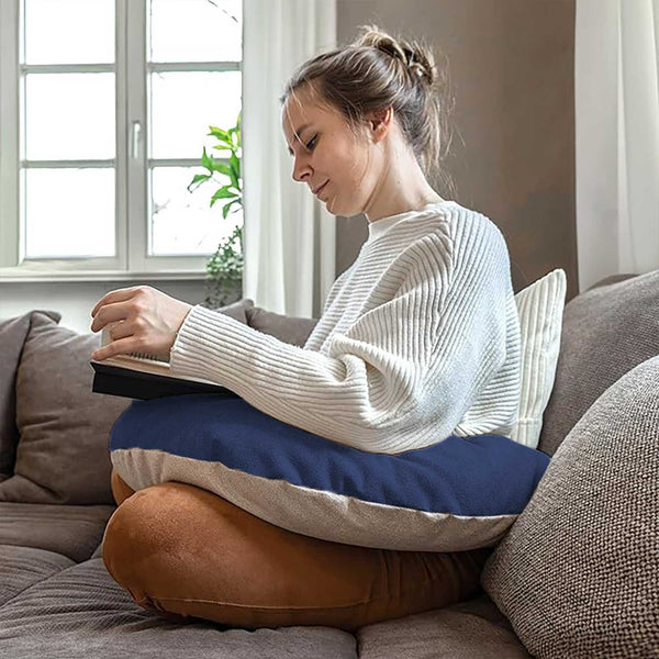 Frau sitzt im Schneidersitz auf einem Sofa und liest ein Buch mit einem großen Kissen auf dem Schoß in einem gemütlichen Raum.
