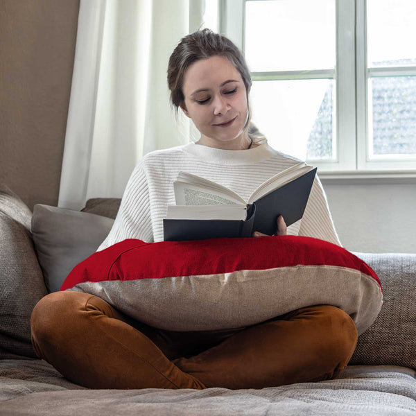 Frau sitzt im Schneidersitz auf einer Couch und liest ein Buch mit einem rot-beigen Kissen auf dem Schoß.