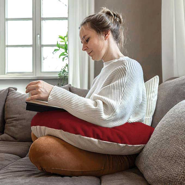 Frau sitzt auf einer Couch, liest ein Buch und hat ein großes rot-beiges Kissen auf dem Schoß.