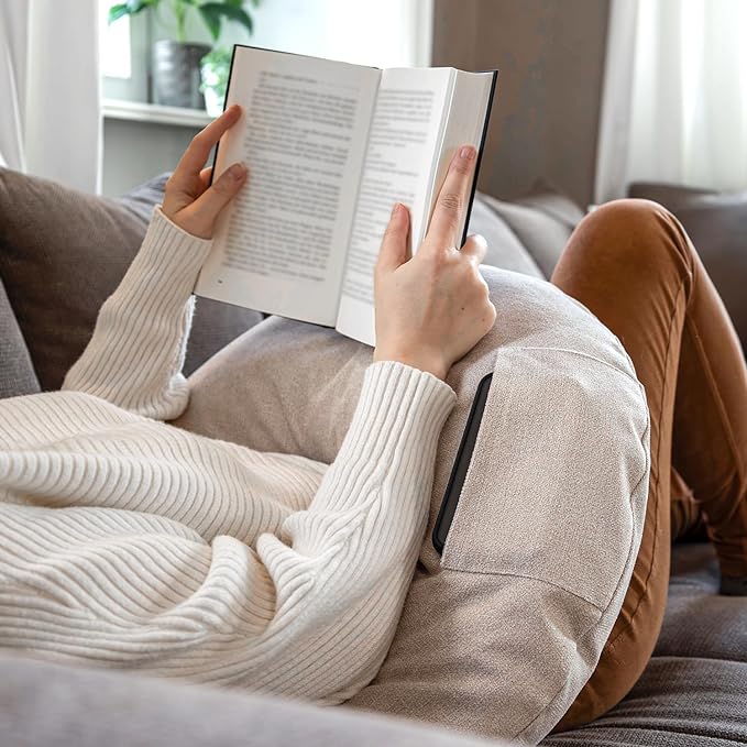 Eine Person entspannt sich auf einem beigen Sofa, liest mit gekreuzten Beinen und einem cremefarbenen Pullover und genießt dank des Lesekissen Classic mit Tasche - Creme stützend im Rücken optimalen Komfort und Langlebigkeit.
