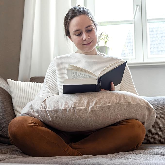 Eine Person sitzt im Schneidersitz auf einem Sofa, hat ein Lesekissen Classic mit Tasche – Creme auf dem Schoß und genießt es, ein Buch zu lesen, während im Hintergrund Sonnenlicht durch das Fenster fällt.