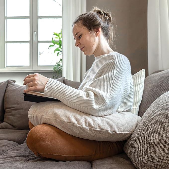 Eine Frau sitzt mit gekreuzten Beinen auf einem Sofa, liest ein Buch und ruht mit den Armen auf dem Lesekissen Classic mit Tasche – Creme, das sie für zusätzlichen Komfort auf dem Schoß hat.