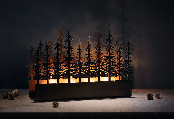 Ein Kerzenhalter aus Metall mit ausgeschnittenen Bäumen, die vom Kerzenlicht beleuchtet werden; in der Nähe sind Tannenzapfen verstreut.