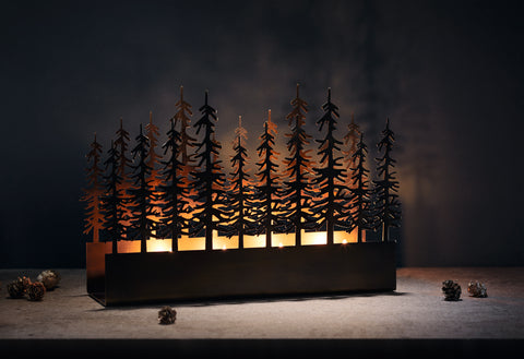 Ein Kerzenhalter aus Metall mit ausgeschnittenen Bäumen, die vom Kerzenlicht beleuchtet werden; in der Nähe sind Tannenzapfen verstreut.