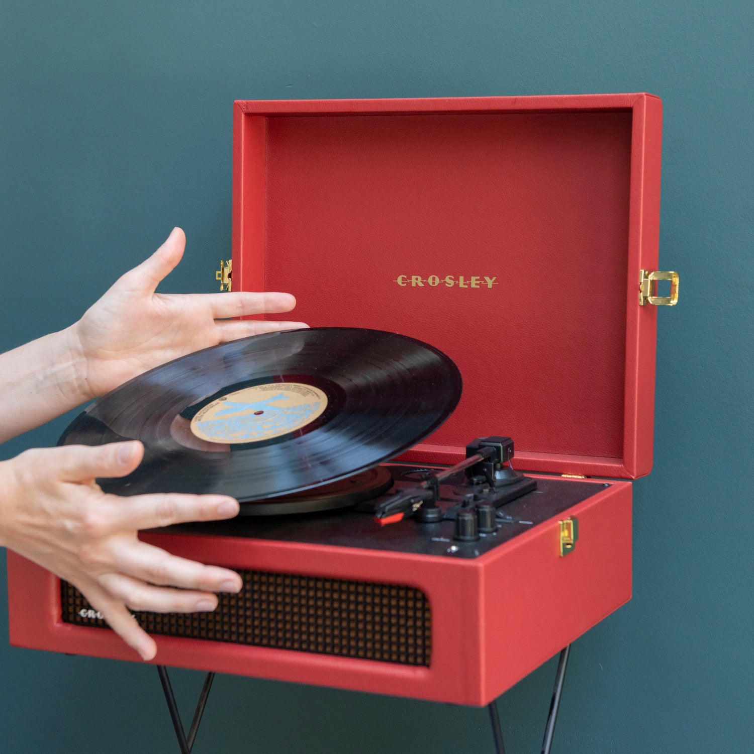 Hände legen eine Schallplatte auf einen roten Crosley-Plattenspieler vor einem blaugrünen Hintergrund.