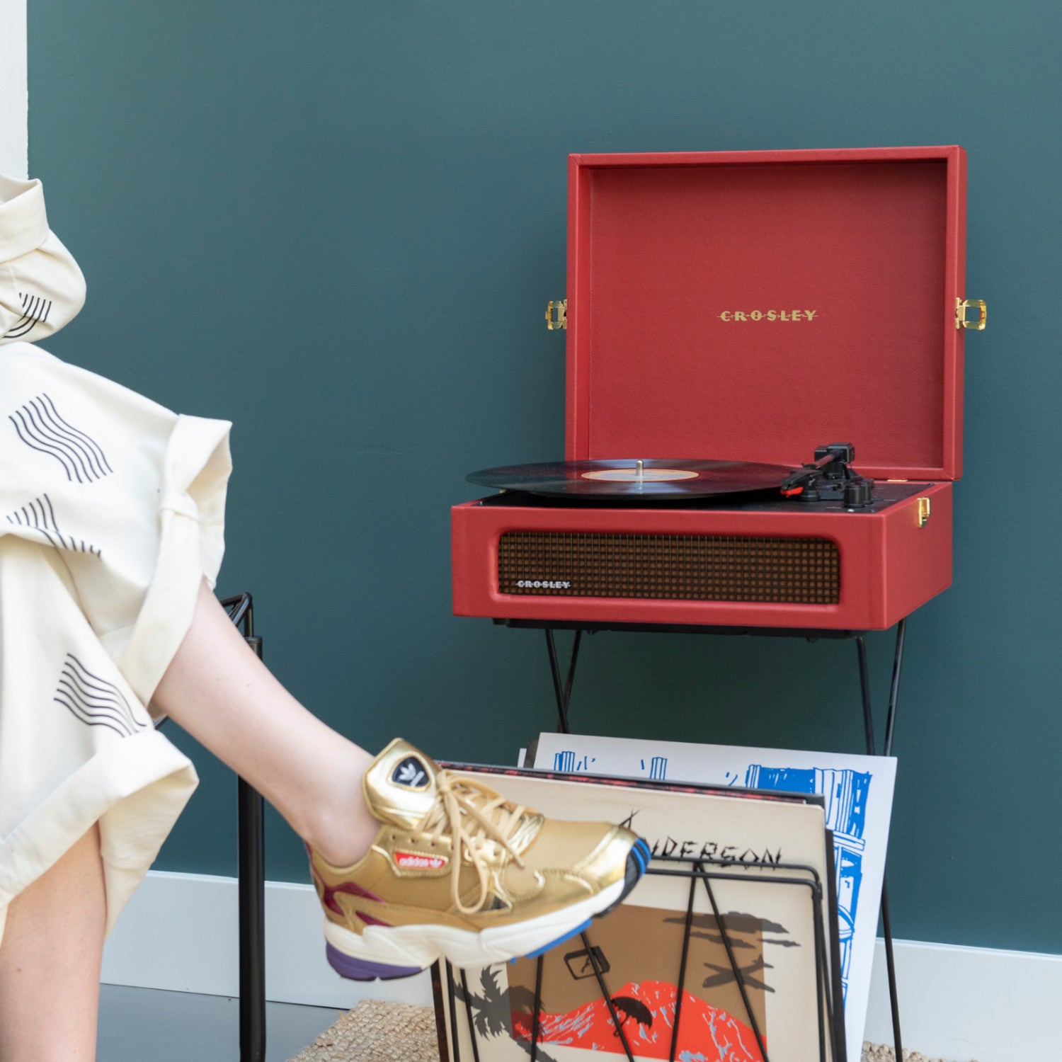 Eine Person in goldenen Turnschuhen sitzt neben einem roten Crosley-Plattenspieler und einem Stapel Schallplatten.