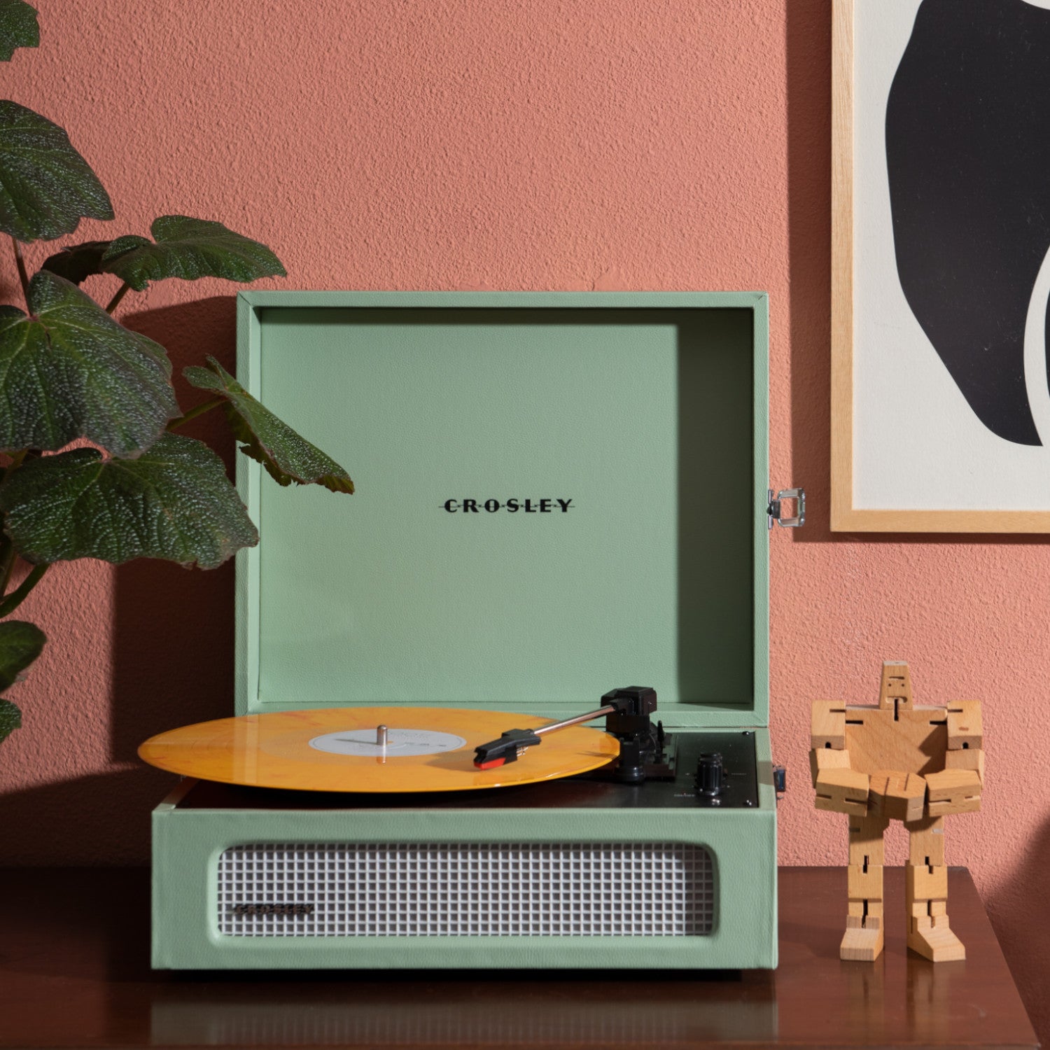 Ein mintgrüner Crosley-Plattenspieler mit gelber Schallplatte steht auf einem Holztisch neben einer hölzernen Roboterfigur.