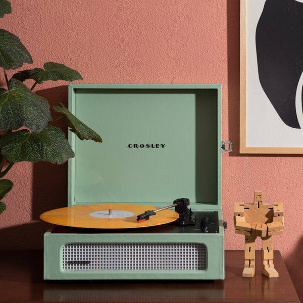 Ein mintgrüner Crosley-Plattenspieler mit gelber Schallplatte steht auf einem Holztisch neben einer hölzernen Roboterfigur.