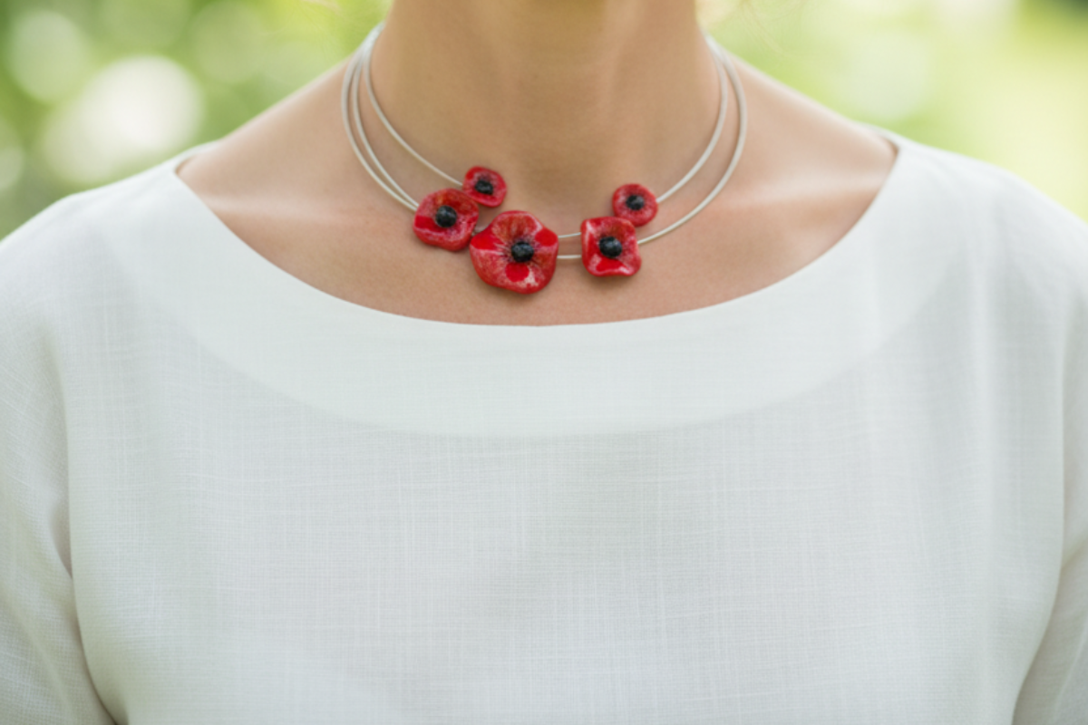 Eine Frau mit hellbraunem Haar, die ein weißes Oberteil und eine Halskette mit roten Blumenanhängern trägt, steht im Freien mit Grünzeug im Hintergrund.
