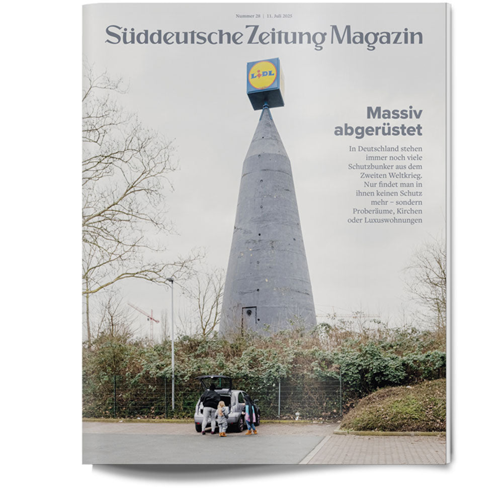 Ein hoher grauer Turm mit einem Lidl-Schild auf der Spitze; davor stehen Menschen an einem offenen Auto, unter einem bewölkten Himmel.