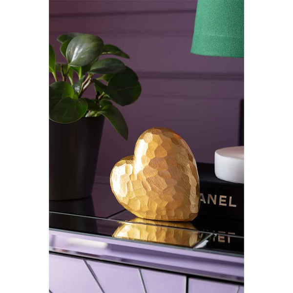 Eine goldene, strukturierte Herzskulptur steht auf einer verspiegelten Fläche neben einer Topfpflanze und gestapelten Büchern.