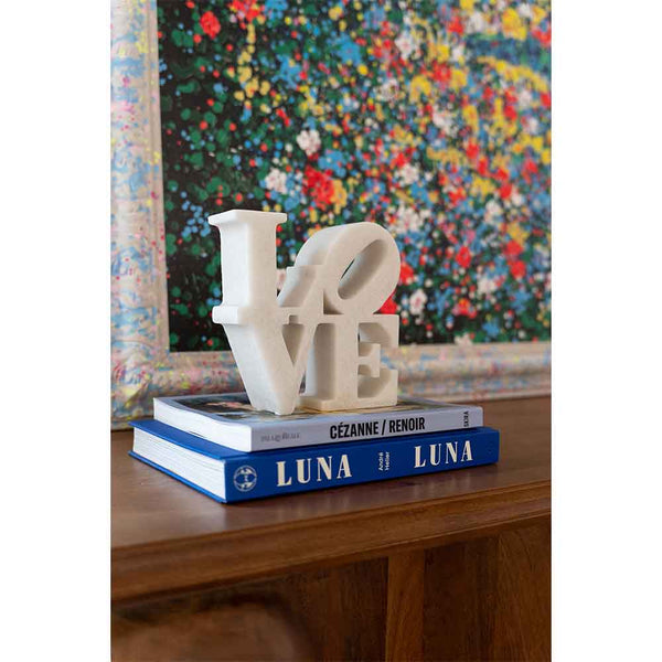 Eine weiße LOVE-Skulptur sitzt auf zwei blauen LUNA-Büchern vor einem bunt gerahmten Gemälde.