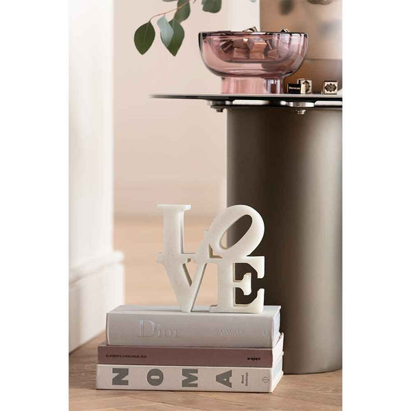 Weiße LOVE-Skulptur auf gestapelten Büchern neben einem modernen Tisch mit einer rosa Glasschale und grünen Blättern darüber.